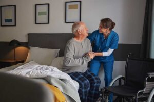 Two seniors and a staff member talking in a bright communal living room with chairs, plants, and activity items visible in the background.
